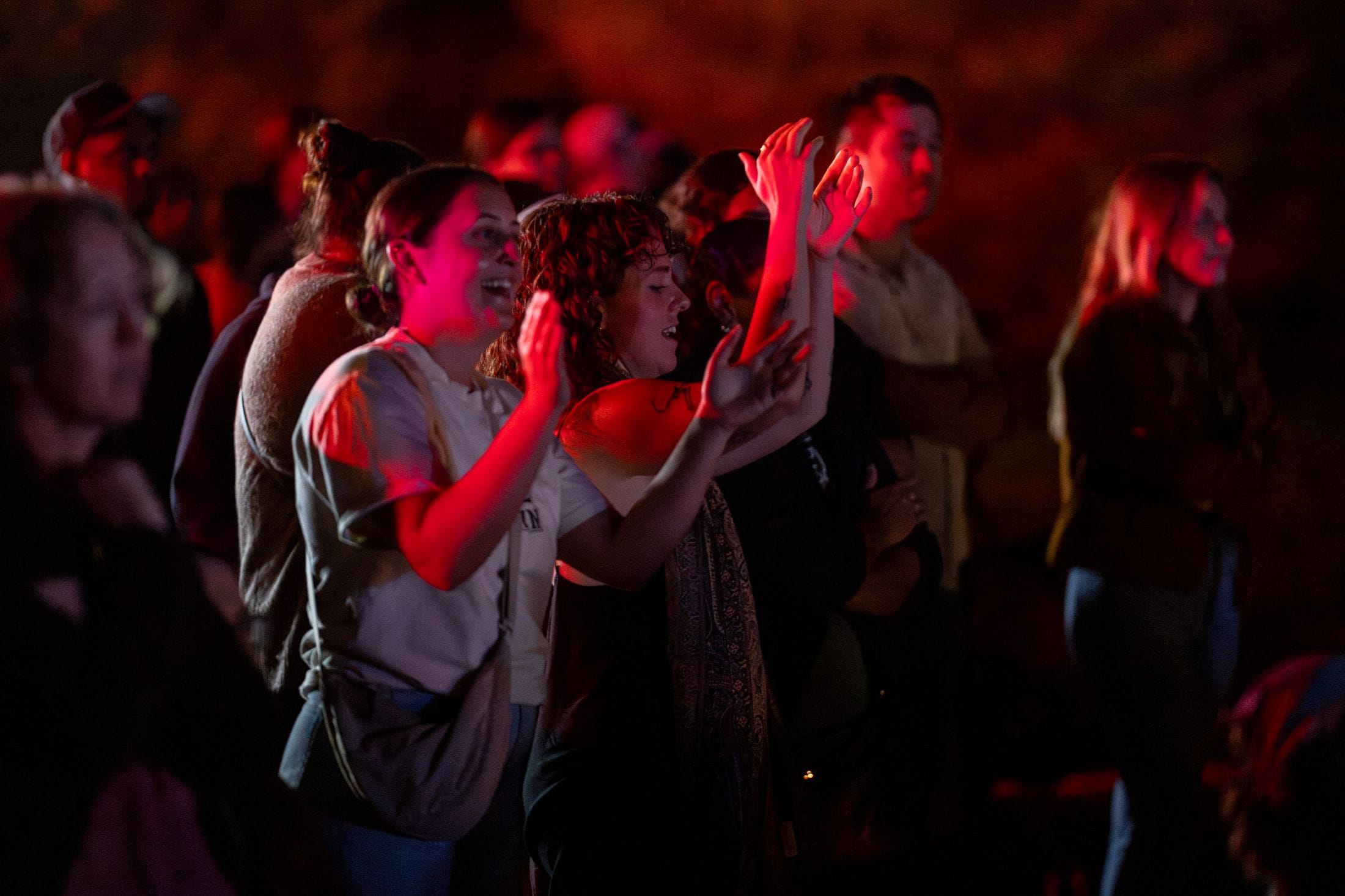 People at a nighttime concert, two audience members in front raising hands and clapping, faces lit by red stage lights.